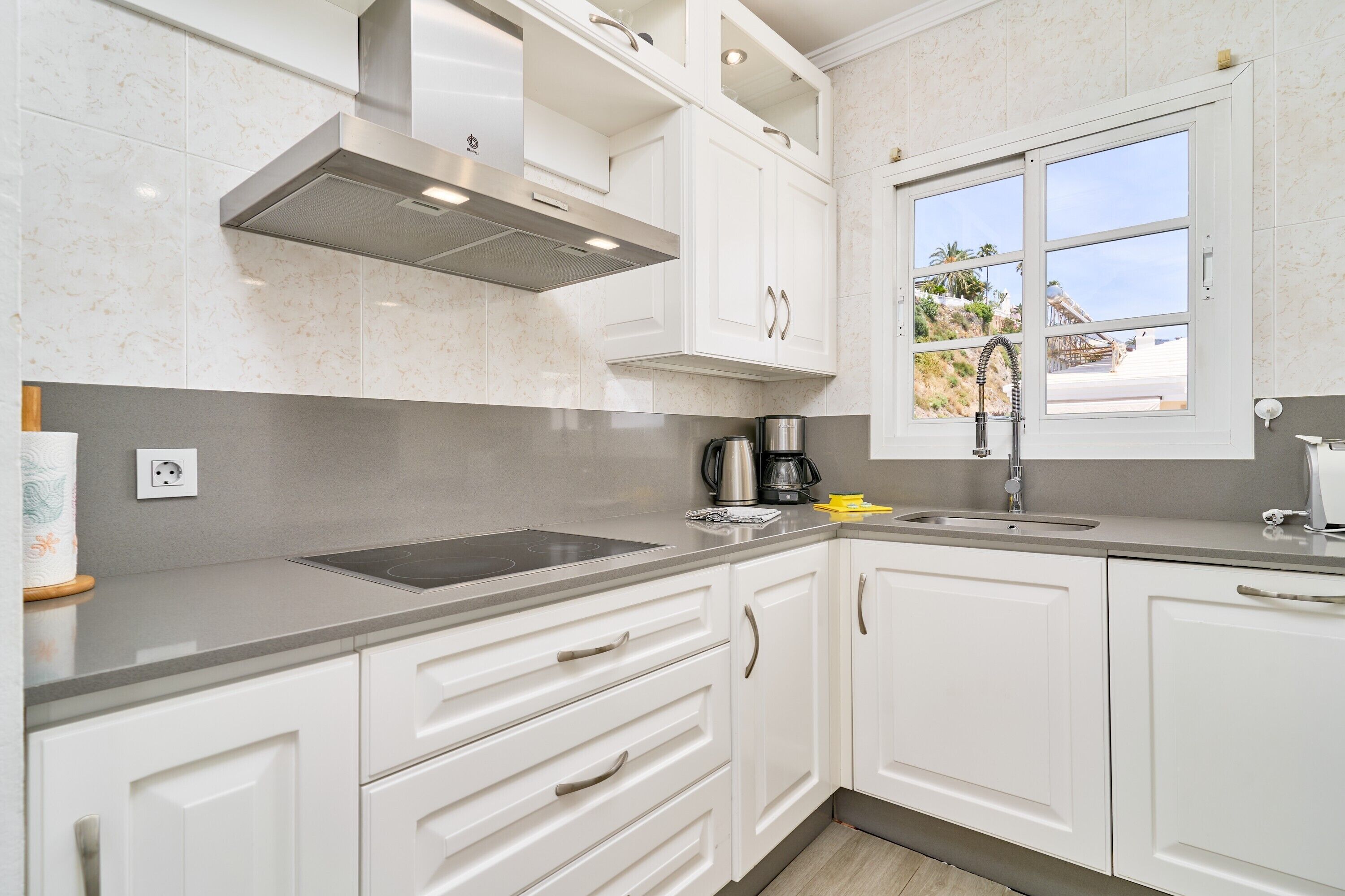 A modern kitchen with white cabinets, a stainless steel range hood, a built-in oven, a sink, and a window with a view of a beach.