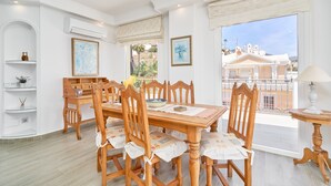 A dining area with a wooden table and chairs, a white shelf with decorative items, and a view of buildings through a window. - Apartment 'Wonderful Penthouse' with Garden, Terrace, Wi-Fi & A/C (Nerja)