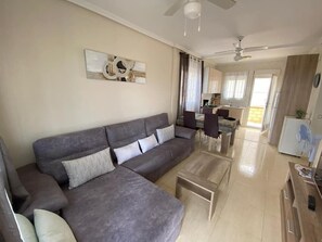 A living room with a grey sofa, a coffee table, a dining area with chairs, and a kitchen in the background.