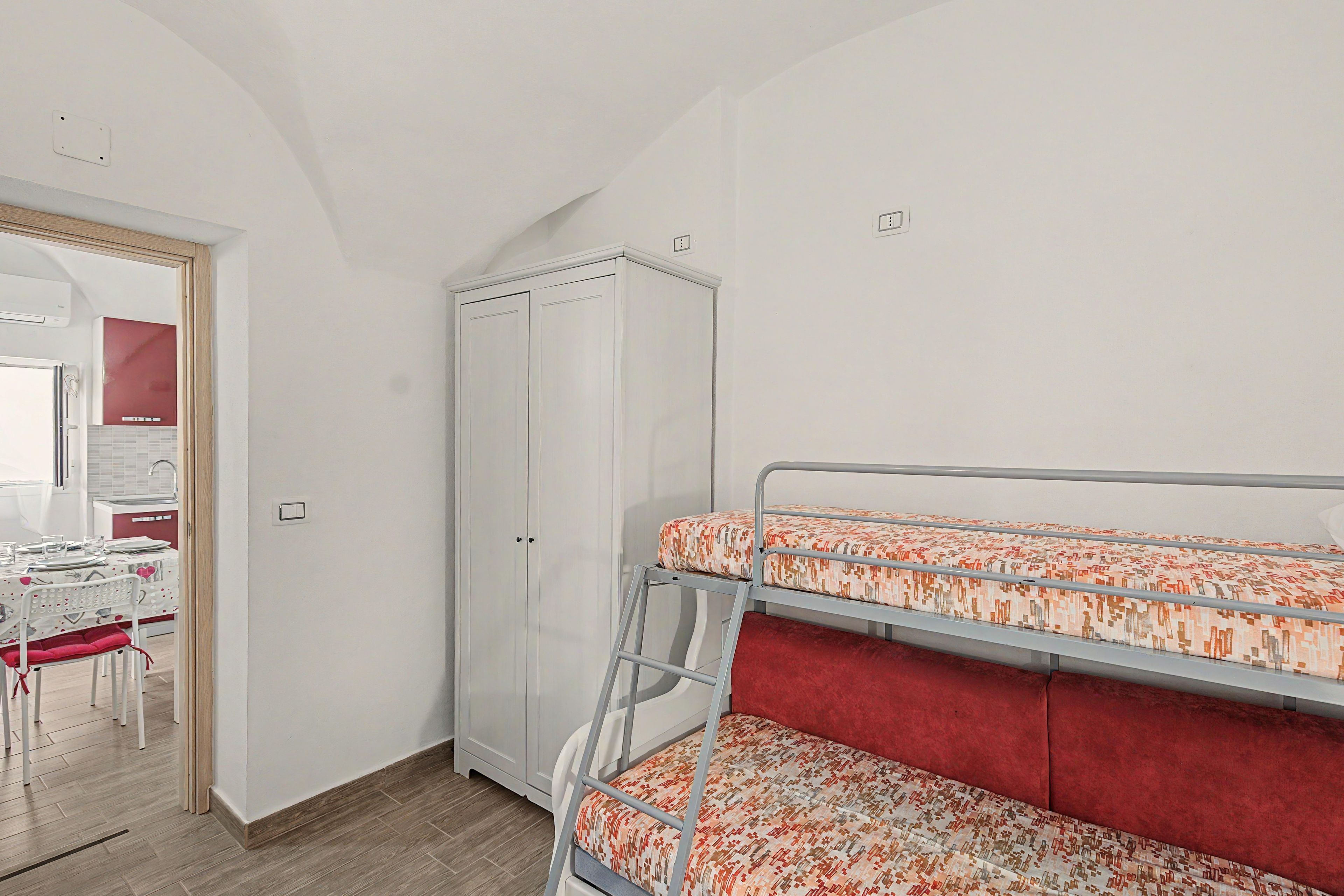 A room with bunk beds, a wardrobe, a desk with chairs, and a kitchen area.