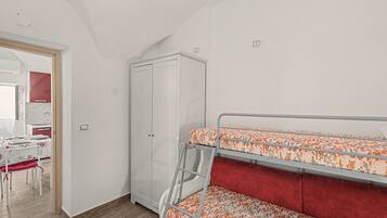 A room with bunk beds, a wardrobe, a desk with chairs, and a kitchen area.