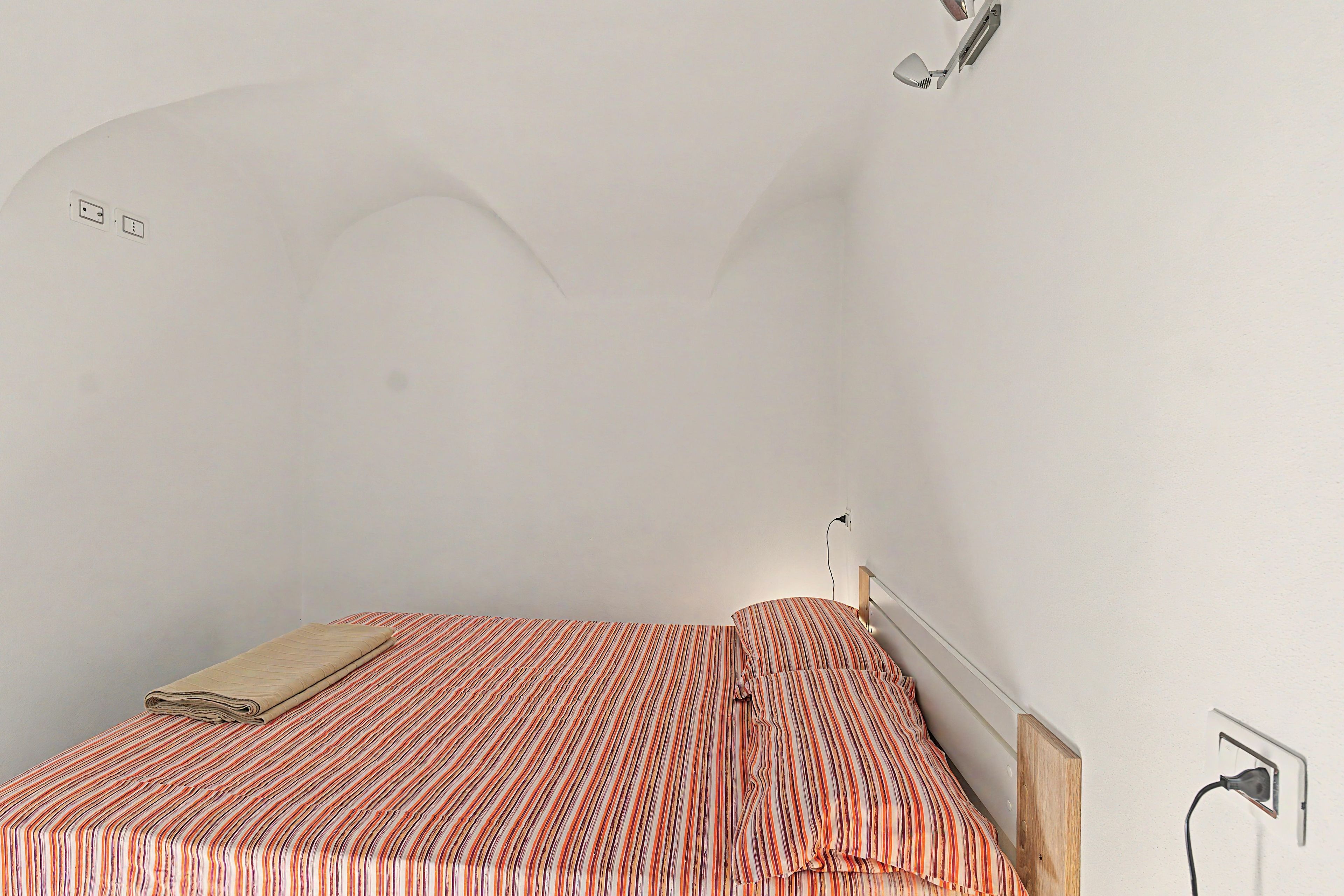 Una camera piccola con un letto singolo, una coperta a righe, una lampada da comodino e una presa elettrica a muro.