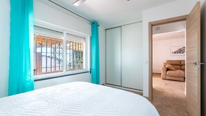 A bedroom with a bed, a ceiling fan, a window with bars, a sliding door, and a sofa in the adjacent room.