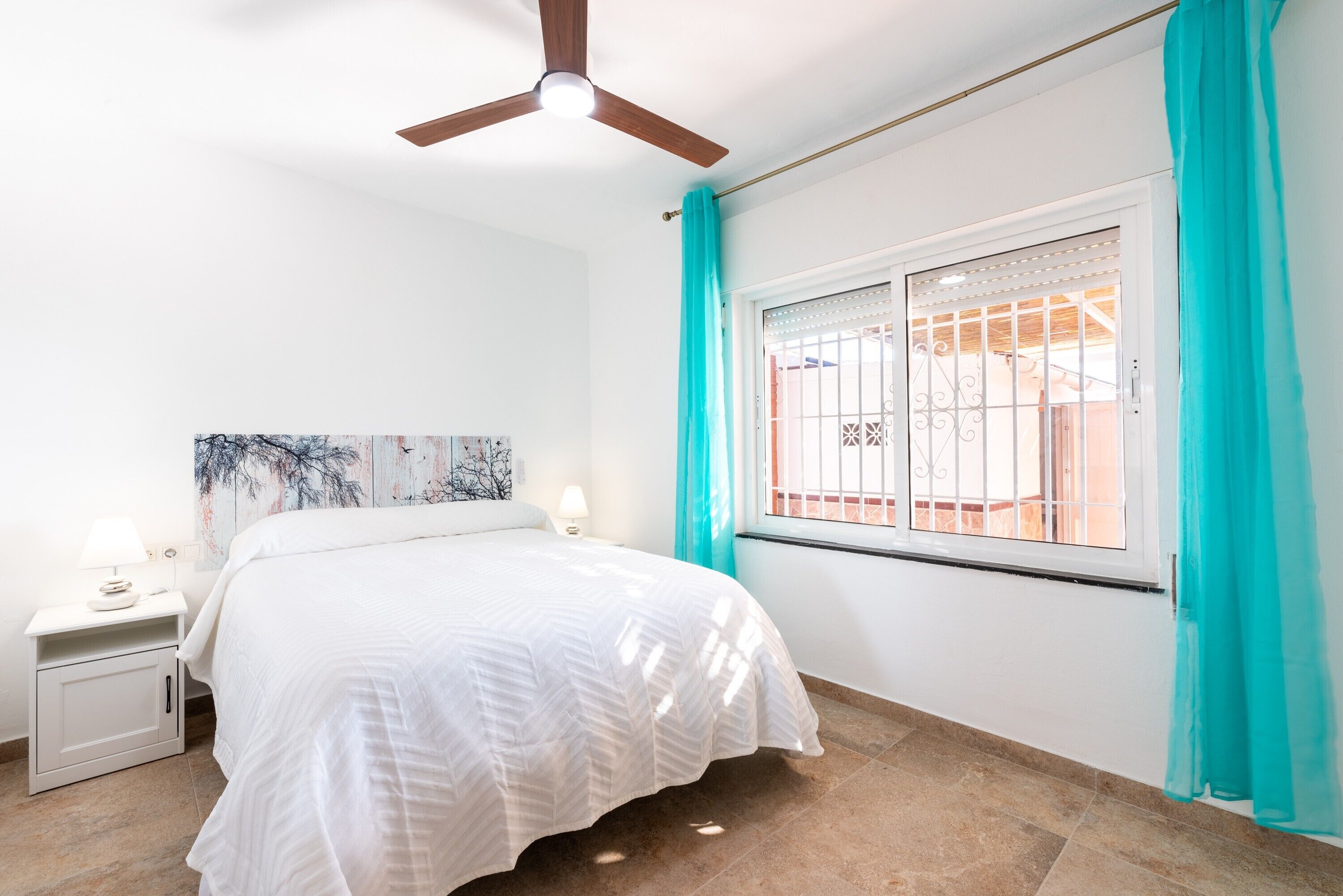 Une chambre à coucher comprenant un lit, une table de chevet, un ventilateur de plafond, une fenêtre avec des rideaux et un luminaire fixé au mur.