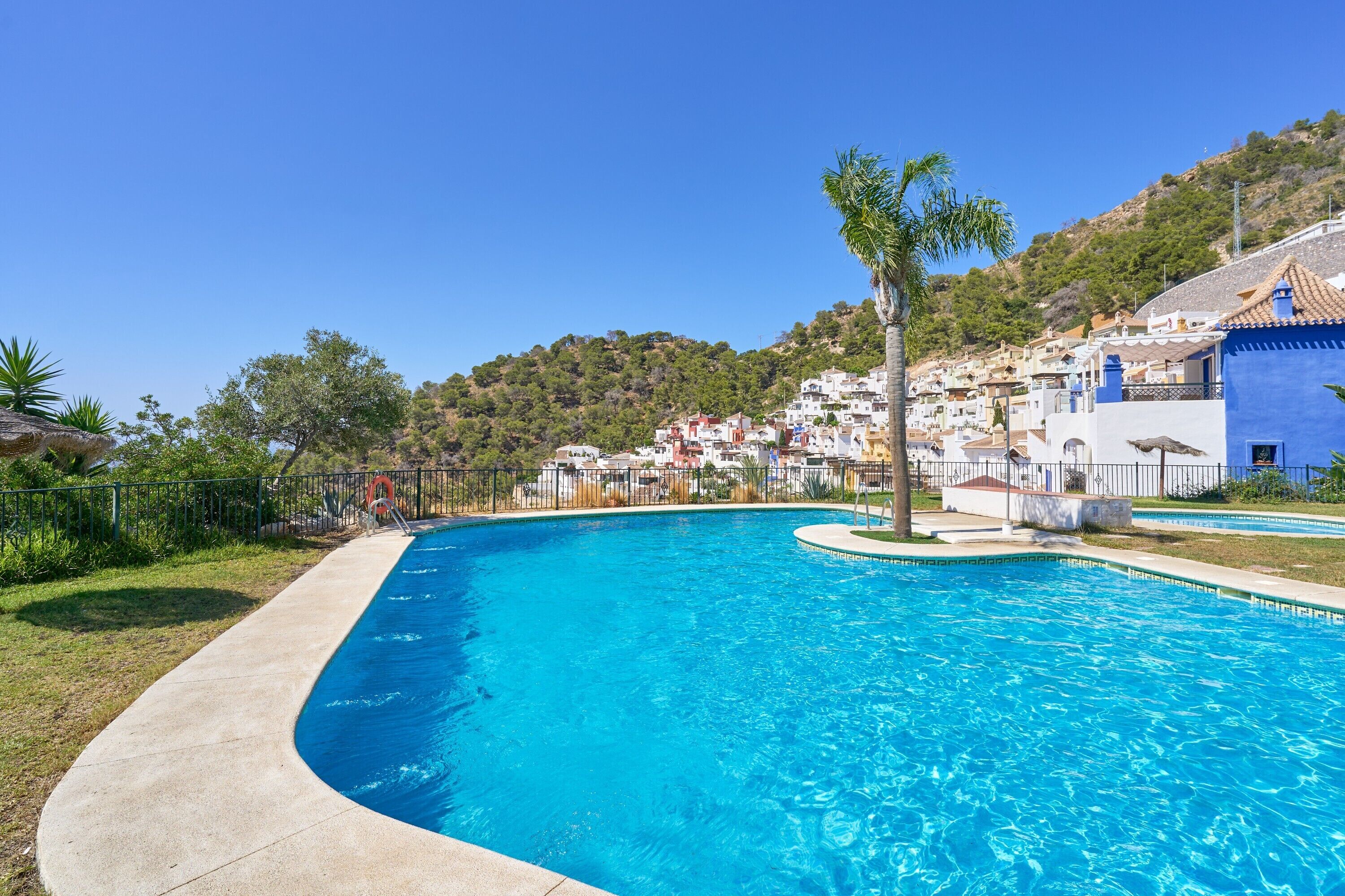A swimming pool with a curved edge, surrounded by a concrete deck. In the background, there are hillside buildings with a mix of colors and a clear blue sky.