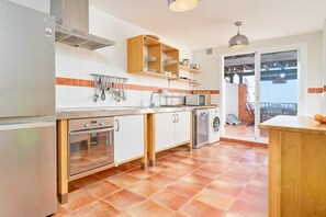 A kitchen with a stainless steel refrigerator, oven, microwave, and washing machine. There are wooden shelves above the counter and a hanging pot rack.