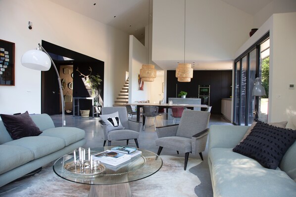 A modern living room with a glass coffee table, grey sofas, and a dining area in the background.