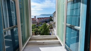 Vista desde una ventana con contraventanas abiertas, que da a un pueblo costero con edificios y cielo despejado.