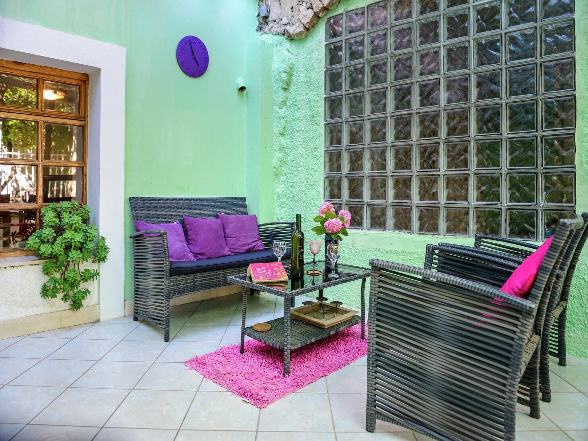 A cozy room with a green wall, a sofa with purple cushions, a coffee table with a vase of flowers, and a window with a view of trees.