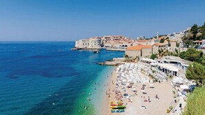 Un pueblo costero con una fortaleza histórica, aguas azul turquesa y una playa concurrida llena de sombrillas y gente.