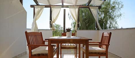 En overdækket terrasse med et træbord og stole, en potteplante og udsigt til træer.
