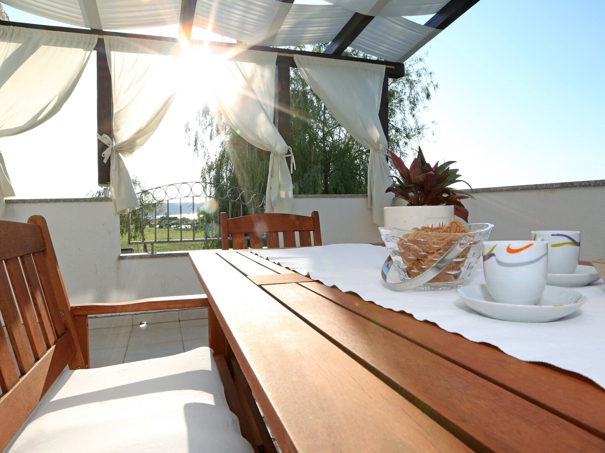 A wooden table with a white tablecloth, a glass bowl of snacks, two white cups with colorful designs, and a potted plant.