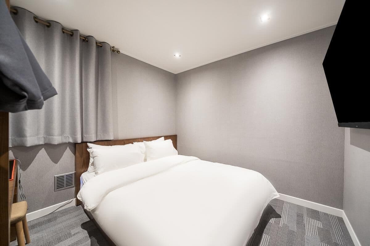 A neatly made bed in a hotel room with a headboard, a flat-screen TV mounted on the wall, and a grey curtain.