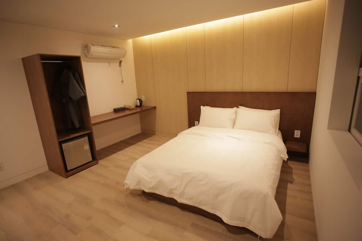 A hotel room with a wooden headboard, a bed with white bedding, a wooden wardrobe, and a small bedside table with a kettle.