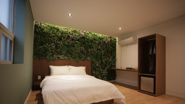 A bedroom with a bed, a wooden headboard, a wall-mounted plant, a wooden wardrobe, and a small desk with a mirror. - Hotel Seven Step (Siheung)