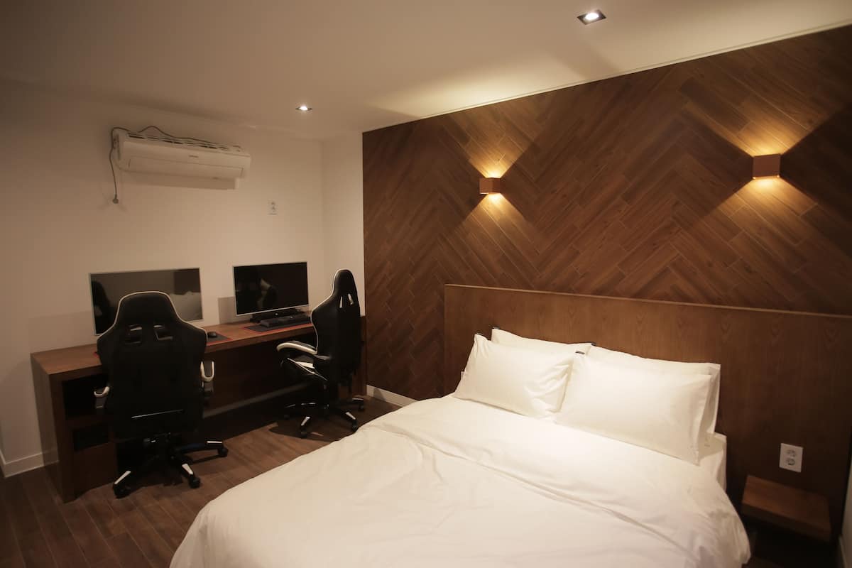 A modern hotel room with a wooden herringbone wall, a bed with white linens, a desk with two monitors, and a black office chair.