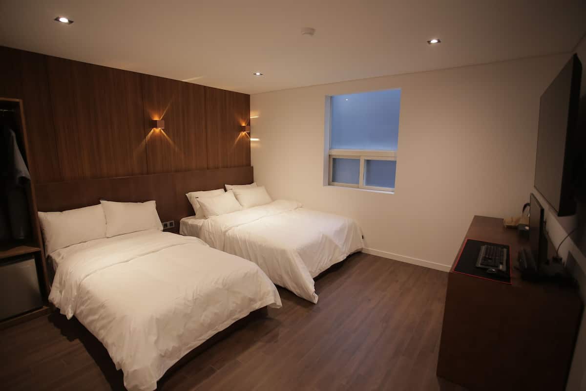 A hotel room with two beds, a wooden headboard, a small window, a desk with a computer, and a TV.