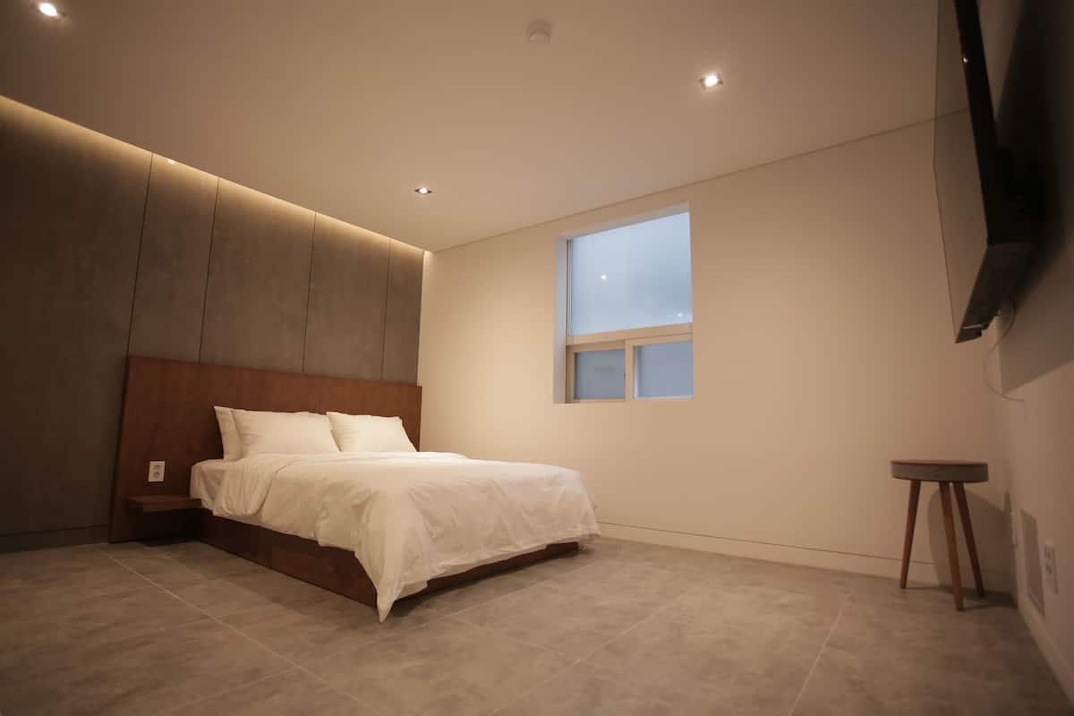 A bedroom with a bed, a small bedside table, a window, and a wall-mounted TV.
