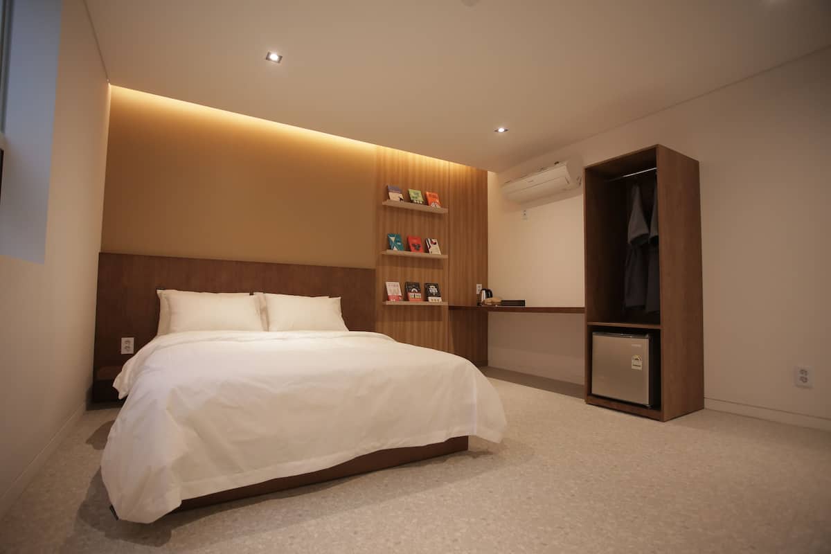 A modern hotel room with a neatly made bed, a wooden headboard, a built-in shelf with decorative items, a wardrobe, and a small refrigerator.