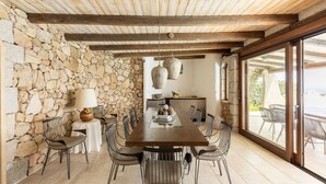 A dining area with a stone wall, wooden ceiling, and large windows. - Luxe Côte Stay- Elegant Villa (Porto Cervo)