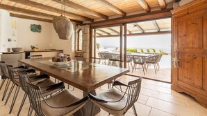A spacious dining area with a large wooden table and chairs, a hanging light fixture, and a view of the sea through large windows. - Luxe Côte Stay- Elegant Villa (Porto Cervo)