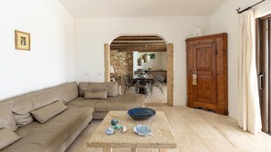 A living room with a beige sofa, a coffee table with decorative items, a wooden cabinet, and a dining area in the background. - Luxe Côte Stay- Elegant Villa (Porto Cervo)