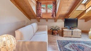 Una habitación acogedora en el ático con techo de madera, un sofá blanco, un televisor sobre un cofre de madera y una planta en maceta.