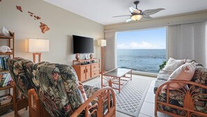 A living room with a television, wooden furniture, a ceiling fan, and a view of the ocean. - Oceanfront Banyan Tree 200 in Kailua Kona, 2BR, 2BA, perfect for 4 guests. (Kailua Kona)