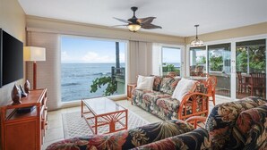 A spacious living room with a large window overlooking the ocean, a ceiling fan, and a dining area with outdoor seating. - Oceanfront Banyan Tree 200 in Kailua Kona, 2BR, 2BA, perfect for 4 guests. (Kailua Kona)