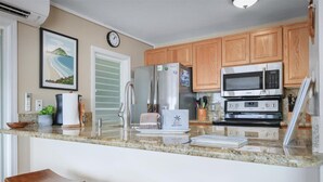 A kitchen with wooden cabinets, stainless steel appliances, a granite countertop, and a clock on the wall. - Oceanfront Banyan Tree 200 in Kailua Kona, 2BR, 2BA, perfect for 4 guests. (Kailua Kona)