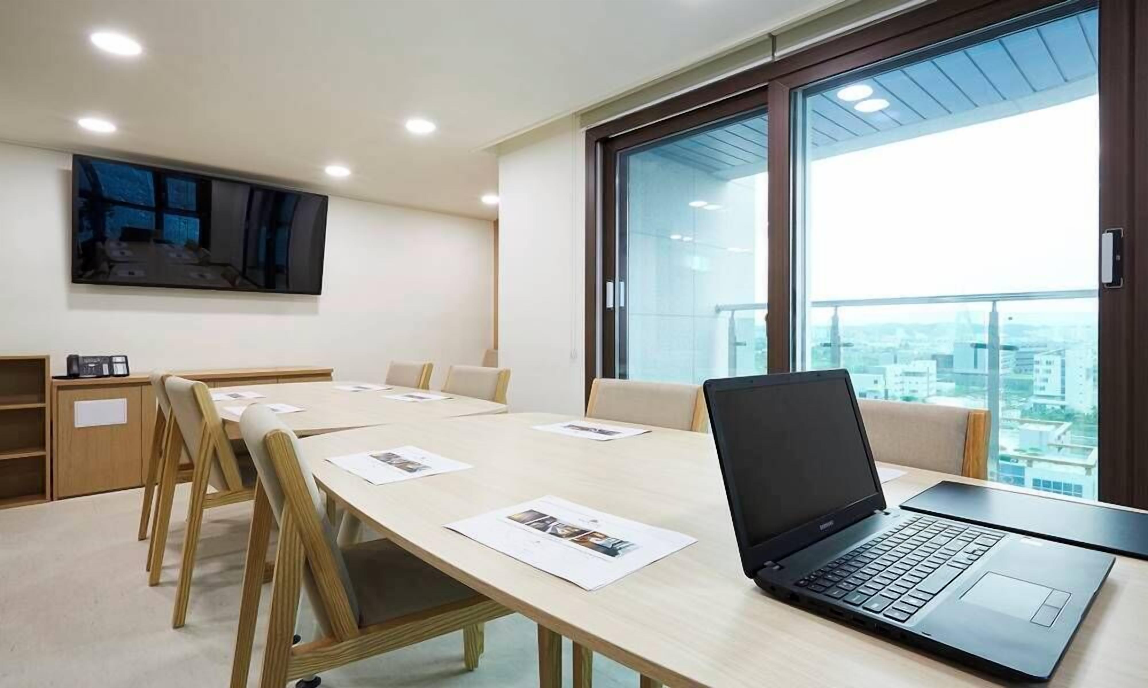 A conference room with a long table, chairs, a laptop, and a large window with a city view.