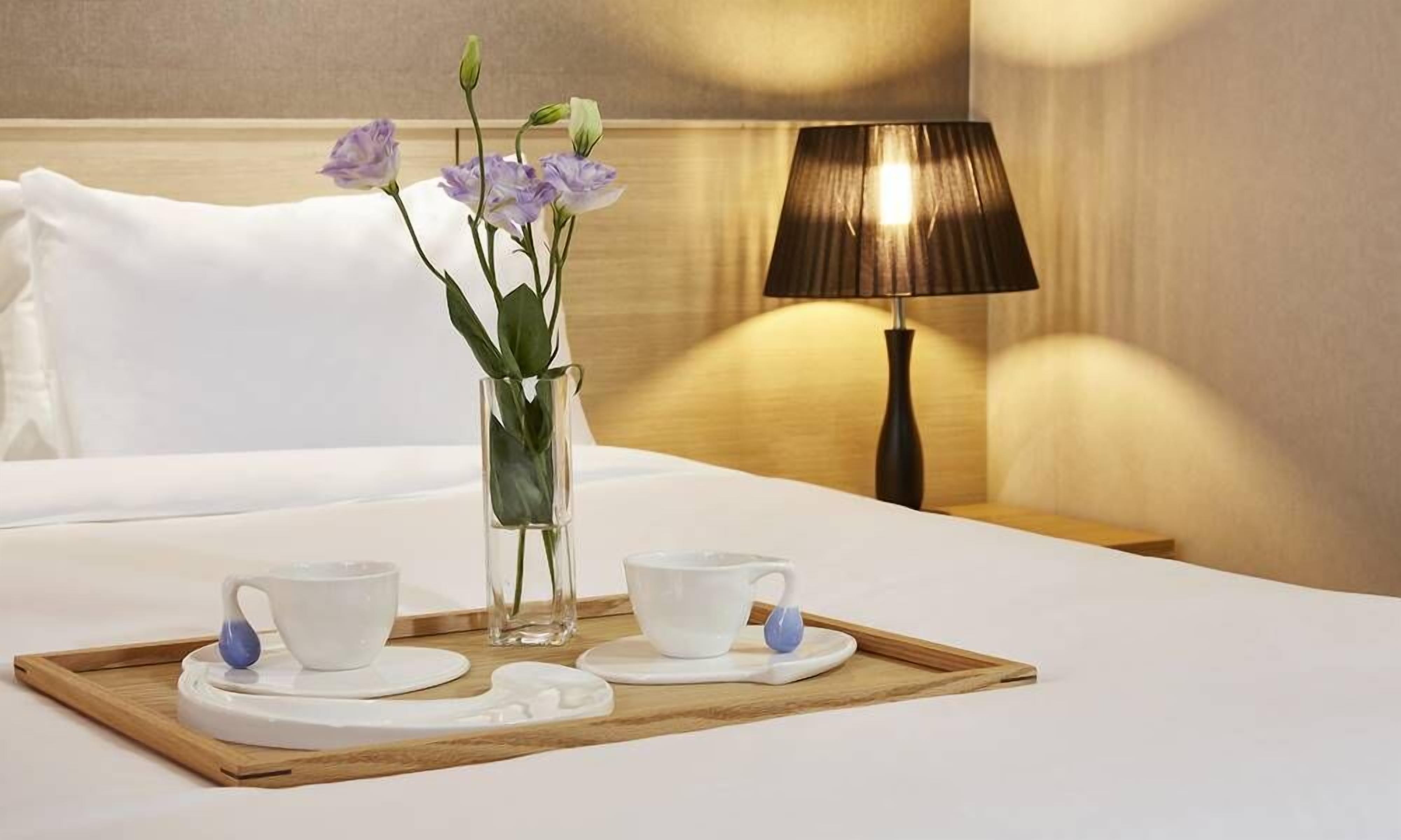 Uma cama bem arrumada com lençóis brancos, um abajur, uma bandeja com duas xícaras e pires brancos, e um vaso com flores roxas.