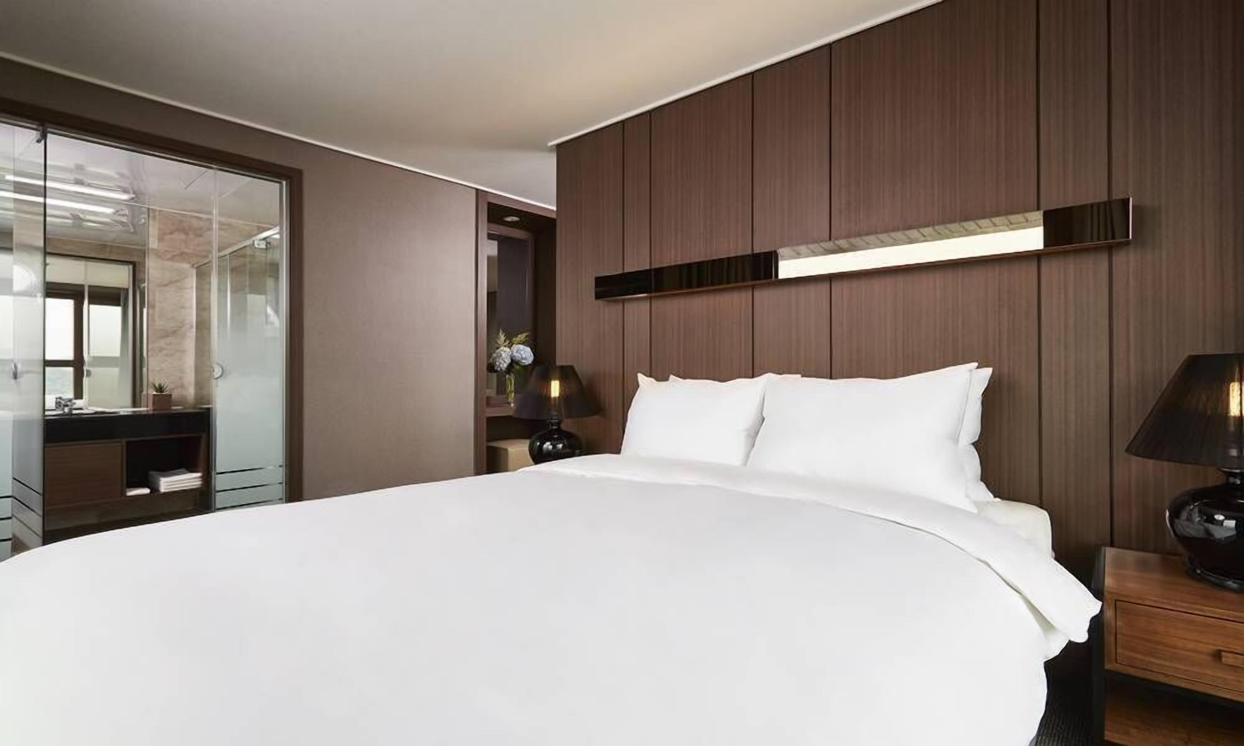 Una habitación de hotel moderna con una cama grande, un panel de pared de madera, una mesita de noche con lámpara y un baño con cabina de ducha de cristal.
