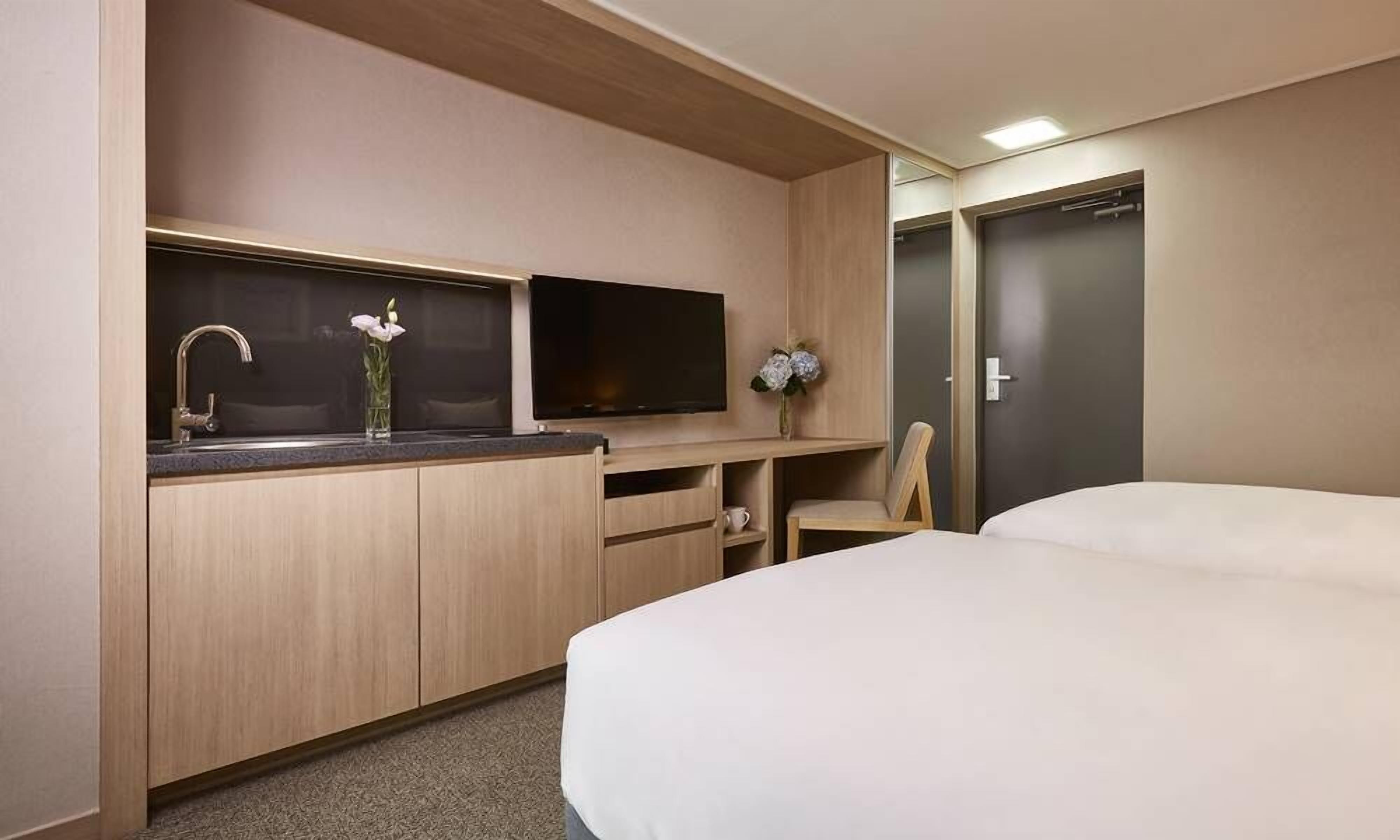 A hotel room with a bed, a TV, a small kitchenette with a sink and a vase of flowers.