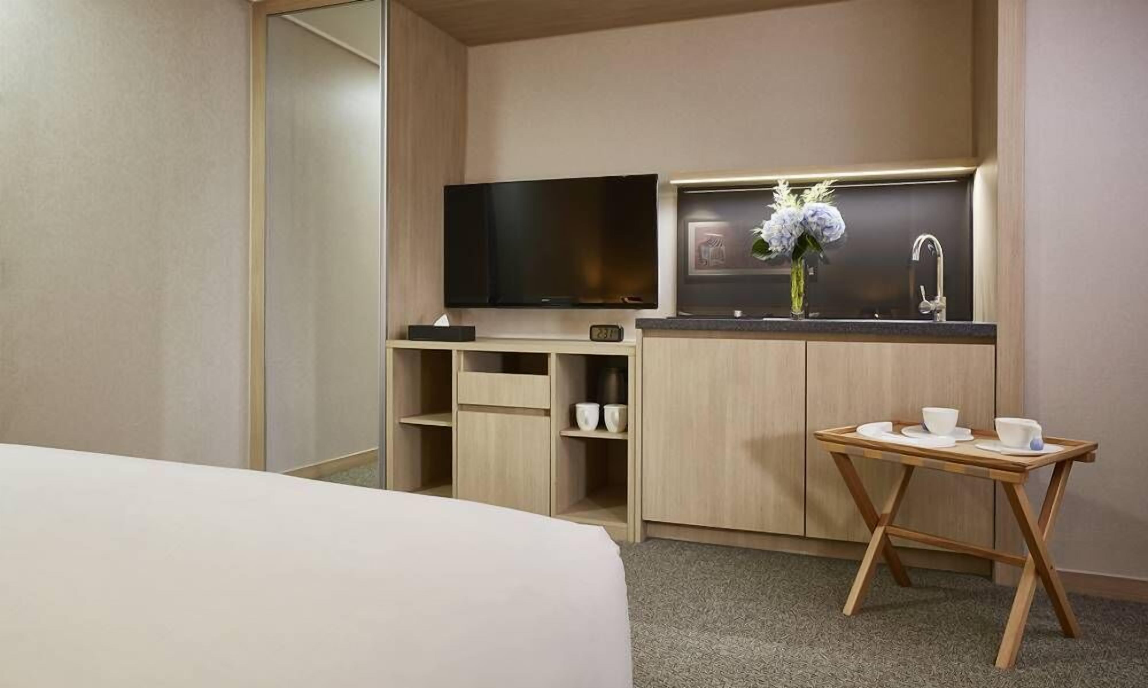 A compact hotel room with a bed, a TV, a small kitchenette with a sink, and a table with cups.