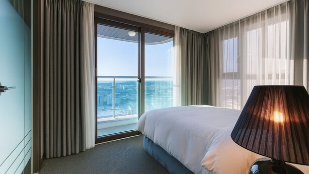 Ein Hotelzimmer mit einem großen Bett, Blick auf die Stadt durch raumhohe Fenster und einem Balkon mit Vorhängen.