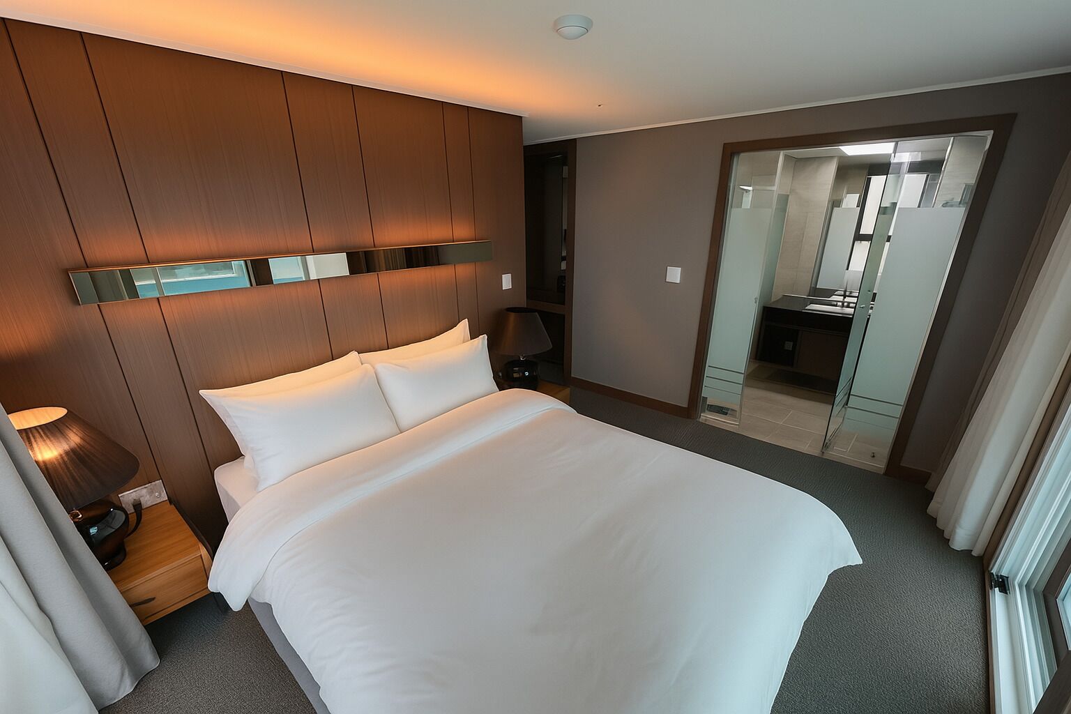 A hotel room with a large bed, a bedside table with a lamp, a mirror, and a view of the bathroom through a glass door.