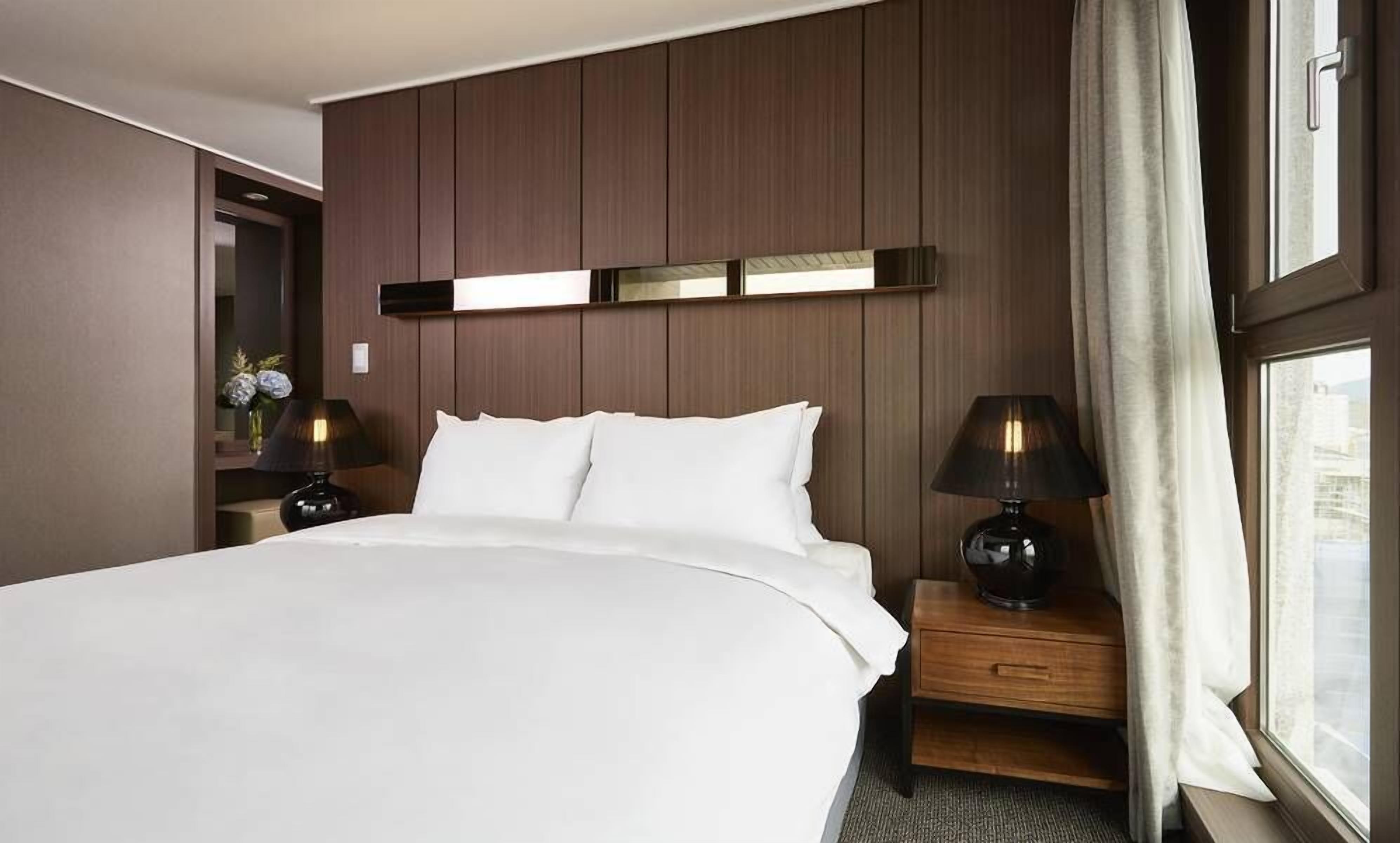 A hotel room with a large bed, wooden headboard, bedside tables with lamps, a mirror, and a window with curtains.