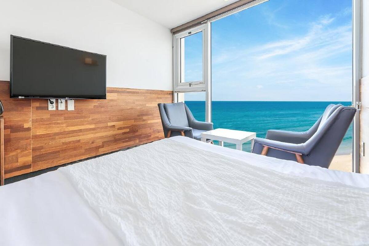 A hotel room with a large bed, a TV mounted on a wooden wall, two chairs, a small table, and a large window offering a view of the ocean.