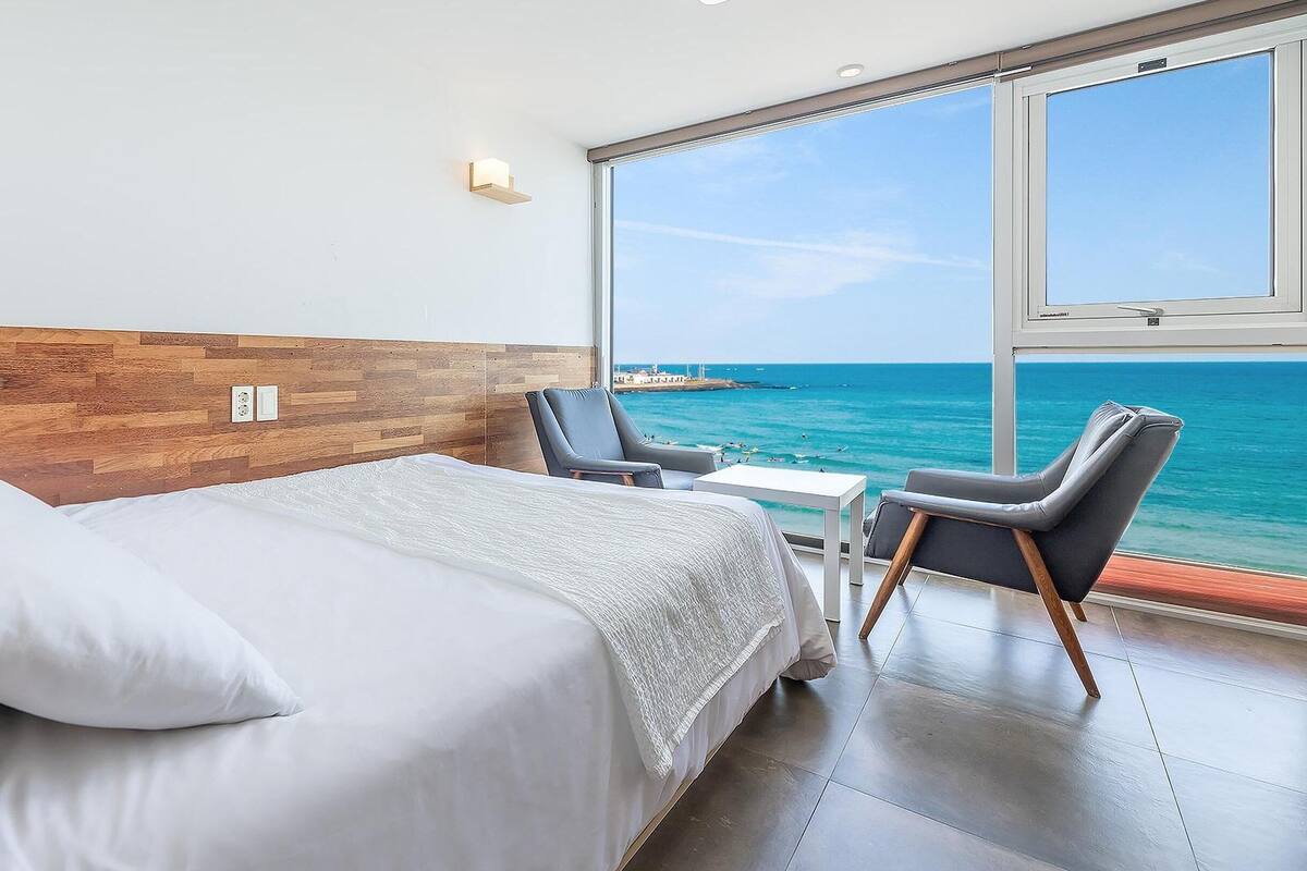 A modern hotel room with a large bed, a small table, two chairs, and a view of the ocean.