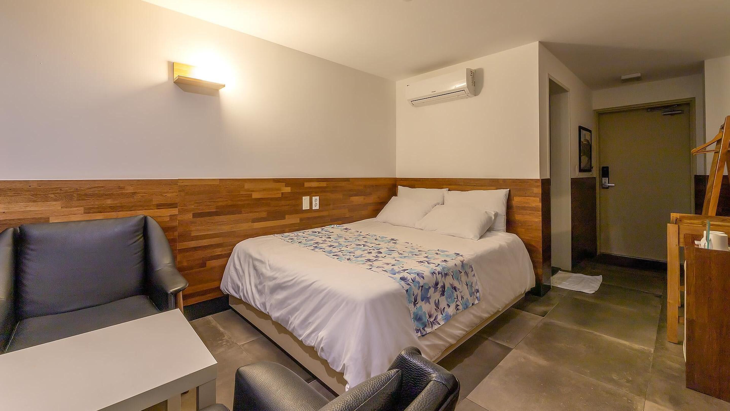 A hotel room with a bed, a desk, a chair, and an air conditioning unit.