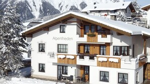 Um edifício de dois andares com o nome 'Alpenfrieden' na fachada, varandas de madeira e vista para montanhas cobertas de neve.