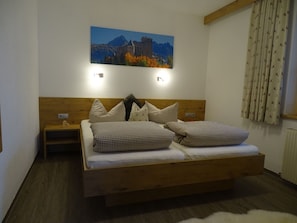 Un dormitorio con cabecera de madera, cama con ropa de cama a cuadros, mesita de noche, una lámpara de pared y un cuadro de un paisaje montañoso.