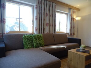 A modern living room with a sectional sofa, a small wooden side table, and a large window with patterned curtains. - Stables - Alpenfrieden (Nauders)