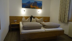Quarto com cabeceira de madeira, cama com roupa de cama xadrez, mesa de cabeceira, luminária de parede e pintura de uma paisagem montanhosa.