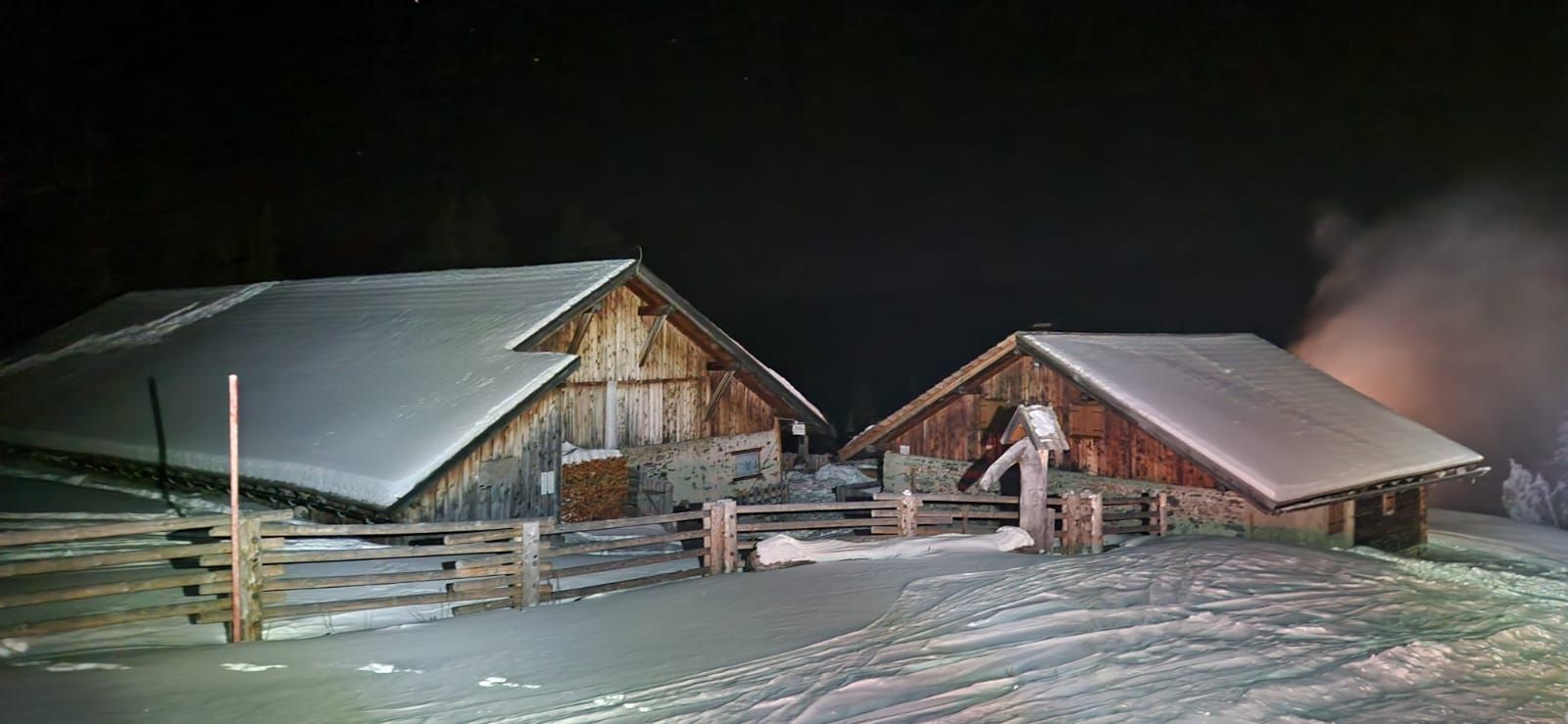 A rustic wooden barn with a metal roof, surrounded by a wooden fence and snow-covered ground.