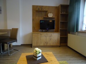 Un salon avec un téléviseur fixé à un mur en bois, un canapé, une petite table ornée d’un objet décoratif et une fenêtre avec des rideaux.