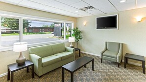 Habitación de hotel con sofá verde, un sillón a juego, una mesita, una lámpara, un televisor montado en la pared y una alfombra estampada.