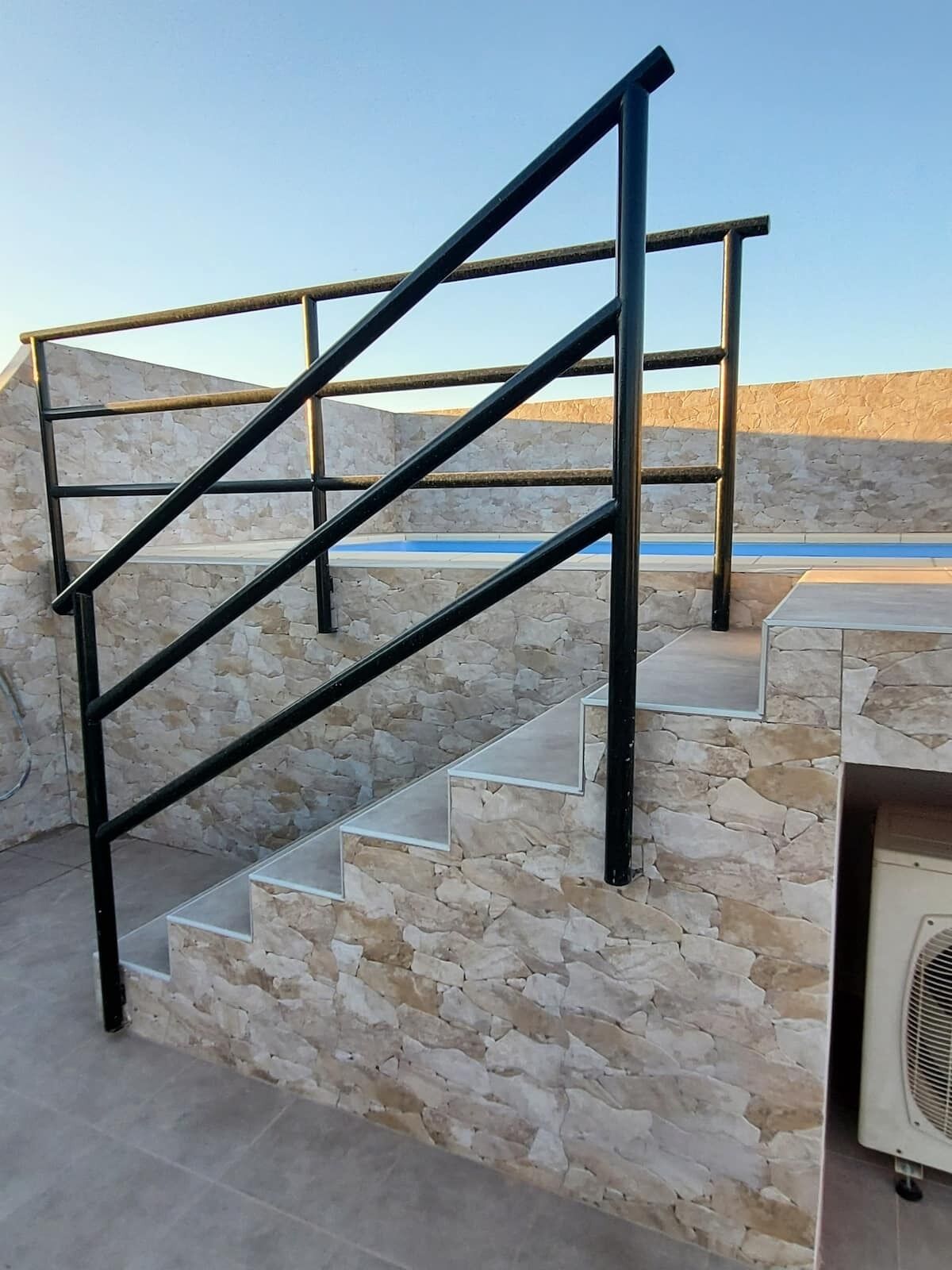 Una escalera de piedra con barandilla de metal que conduce a una terraza con muro de piedra y una piscina al fondo.