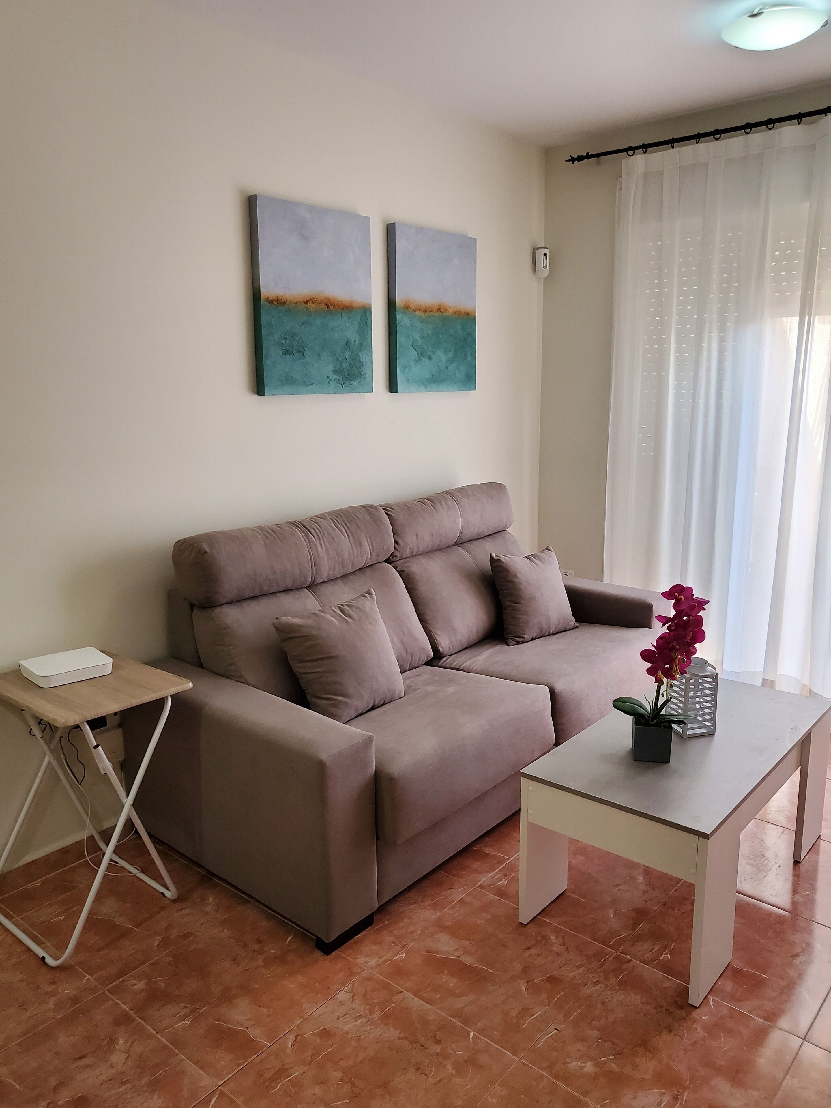 Un salon avec un canapé beige, une petite table d’appoint en bois sur laquelle se trouve un appareil blanc, une table basse blanche avec un pot de plante, et deux tableaux abstraits accrochés au mur.